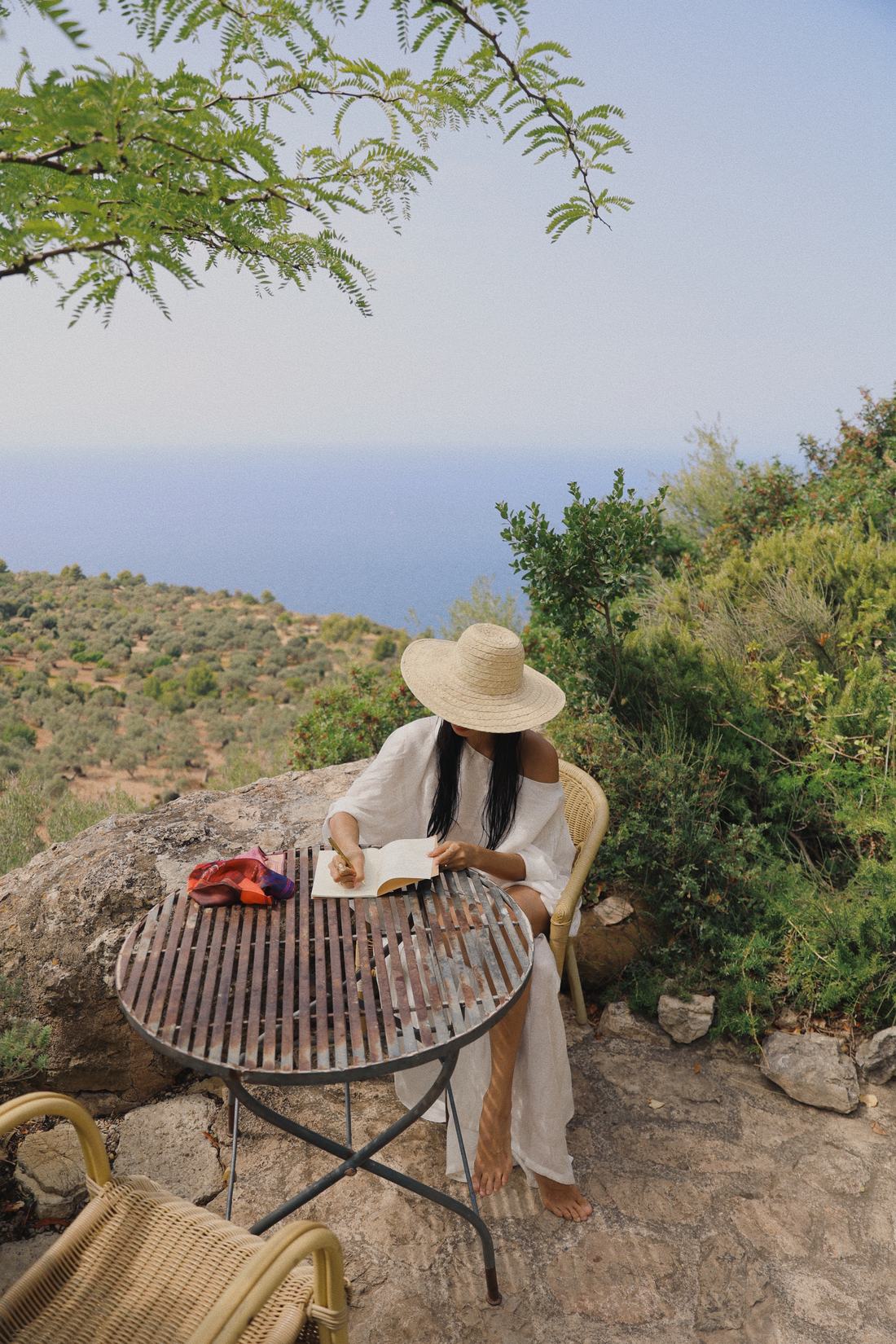 Writing in a notebook at an iron table on a stone terrace above the sea