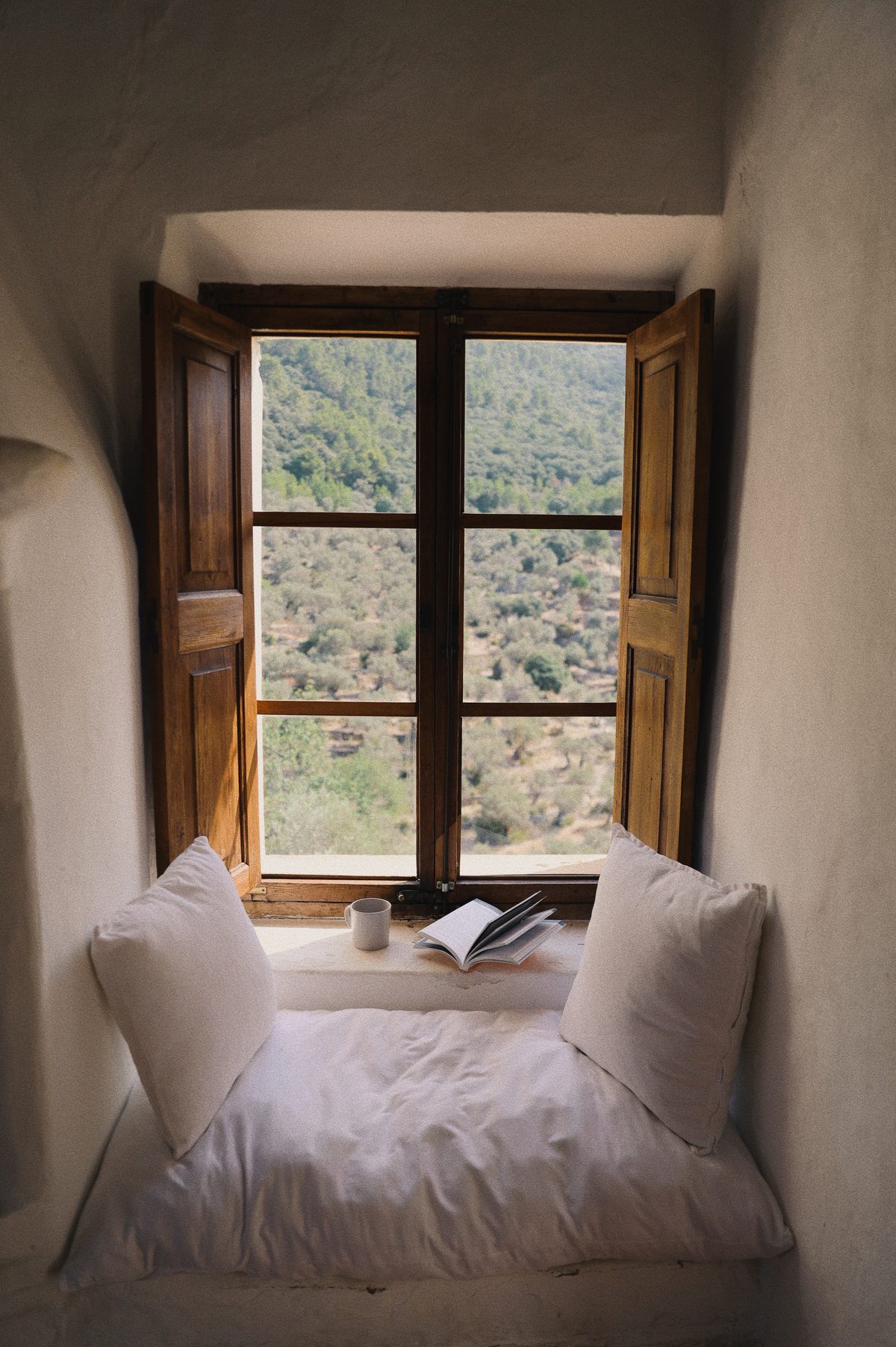 A window seat with white cushions, wooden shutters open to olive hills